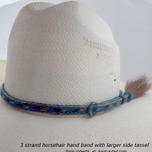 May include: A white cowboy hat with a turquoise, brown, and black horsehair hand band and a larger side tassel. The band is braided and the tassel is made of horsehair. The text "3 strand horsehair hand band with larger side tassel from roberta at knot-a-tail.com" is at the bottom of the image.