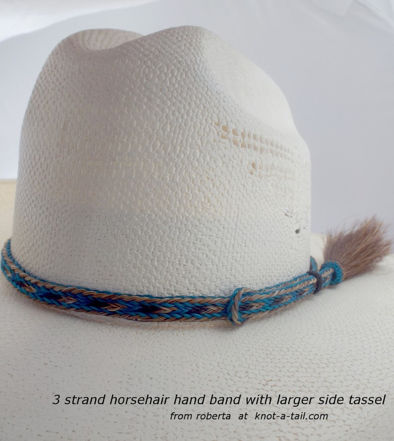 May include: A white cowboy hat with a turquoise, brown, and black horsehair hand band and a larger side tassel. The band is braided and the tassel is made of horsehair. The text "3 strand horsehair hand band with larger side tassel from roberta at knot-a-tail.com" is at the bottom of the image.