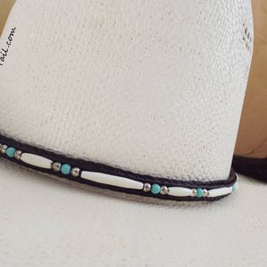 May include: A white cowboy hat with a black braided hat band accented with turquoise and white beads. The hat band has the text "knot-a-tail.com" on it.