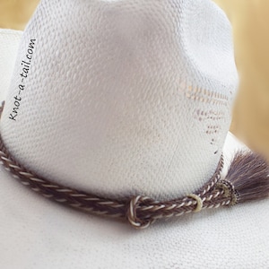 May include: A white cowboy hat with a brown and white braided leather hat band with a brown horsehair tassel. The hat band has the text "knot-a-tail.com" on it.