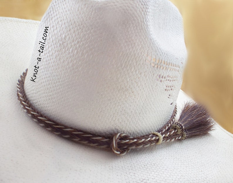 May include: A white cowboy hat with a brown and white braided leather hat band with a brown horsehair tassel. The hat band has the text "knot-a-tail.com" on it.