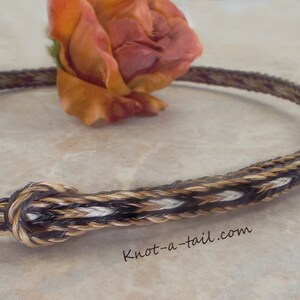 Puede incluir: Un accesorio trenzado para cola de caballo con cierre de nudo. El accesorio presenta una mezcla de hebras marrones, beige y blancas. El texto "knot-a-tail.com" es visible. Una rosa naranja seca est&aacute; en el fondo.