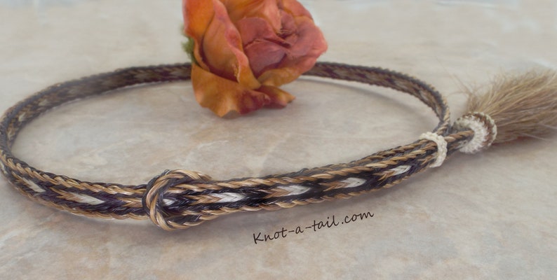 Puede incluir: Un accesorio trenzado para cola de caballo con cierre de nudo. El accesorio presenta una mezcla de hebras marrones, beige y blancas. El texto "knot-a-tail.com" es visible. Una rosa naranja seca est&aacute; en el fondo.