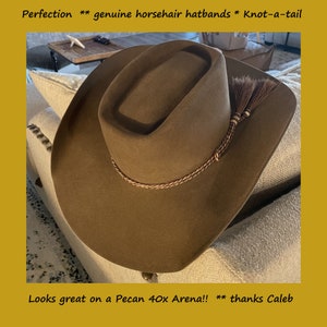 May include: A brown cowboy hat with a horsehair hatband and a knotted tail. The hatband is braided with brown and white thread. The hat is sitting on a light brown couch.