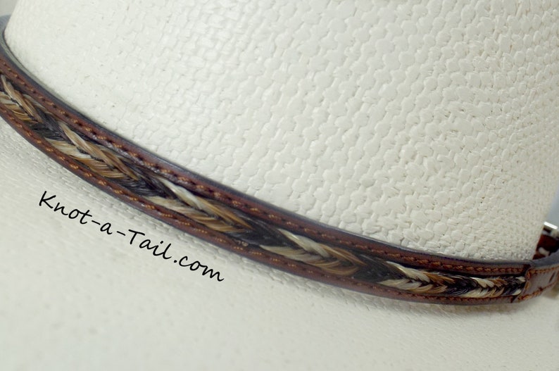 May include: A close-up of a brown leather hat band with a braided horsehair design. The band is on a white hat. The text "Knot-a-Tail.com" is visible on the hat.