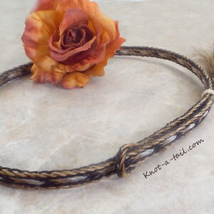 Puede incluir: Collar trenzado de crin de caballo con un patr&oacute;n marr&oacute;n, beige y blanco. El collar est&aacute; anudado y tiene una peque&ntilde;a cola en el extremo. Una rosa naranja y amarilla se coloca cerca del collar. El sitio web knot-a-tail.com es visible.