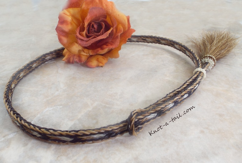 Puede incluir: Collar trenzado de crin de caballo con un patr&oacute;n marr&oacute;n, beige y blanco. El collar est&aacute; anudado y tiene una peque&ntilde;a cola en el extremo. Una rosa naranja y amarilla se coloca cerca del collar. El sitio web knot-a-tail.com es visible.