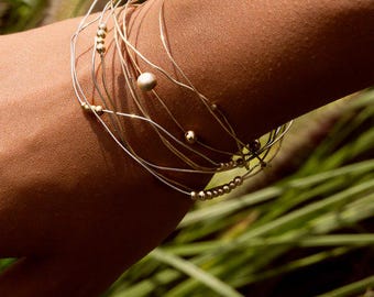 May include: A set of layered bracelets with fine, silver-coloured wires and small, gold-coloured beads. One bracelet has a larger, round, beige bead. The bracelets are worn on an arm, with green foliage in the background.