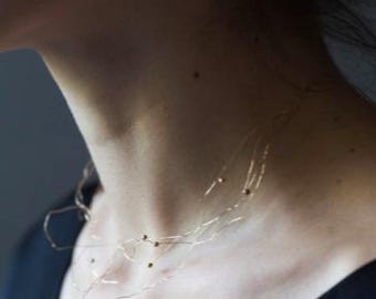 May include: Delicate gold-toned necklace with small, dark beads. The necklace has a unique, organic design, appearing to float against the skin. The jewelry is worn against a dark background, highlighting the necklace's intricate details. A minimalist piece.