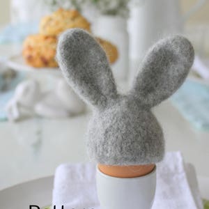 May include: A grey felt bunny rabbit egg topper sits on top of a white egg cup. The egg cup is on a white napkin and a green and white patterned plate. The word "Pattern" is written in black text on the plate.