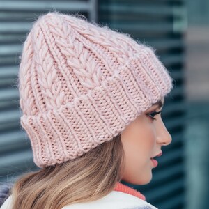 May include: A pink knitted beanie with a cable knit pattern. The beanie is worn by a person with long, light brown hair.