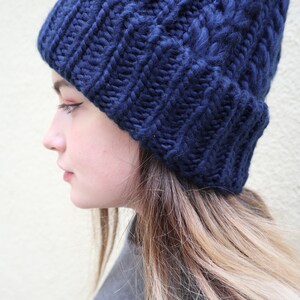 May include: A navy blue knitted beanie with a cable knit pattern. The beanie is worn by a person with long blonde hair.