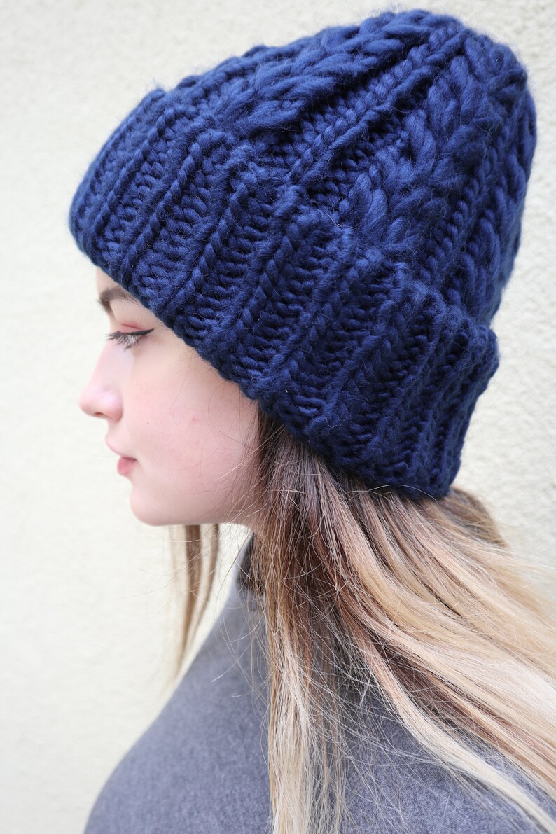 May include: A navy blue knitted beanie with a cable knit pattern. The beanie is worn by a person with long blonde hair.