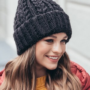 May include: A black knitted beanie with a cable knit pattern. The beanie is worn by a person with long brown hair.