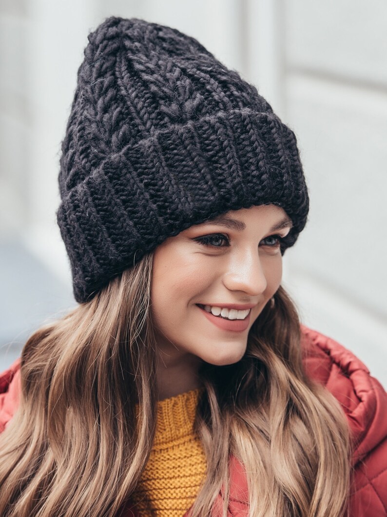 May include: A black knitted beanie with a cable knit pattern. The beanie is worn by a person with long brown hair.