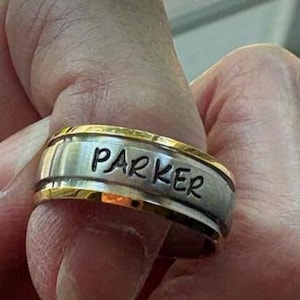 May include: A two-tone metal ring featuring a brushed silver-colored center band with the name PARKER engraved in black capital letters, bordered by polished gold-colored edges on the top and bottom of the band.