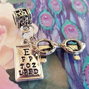 May include: Silver charm with a silver eye chart pendant and a pair of silver glasses charm. The eye chart says "EFPTOZLPED".