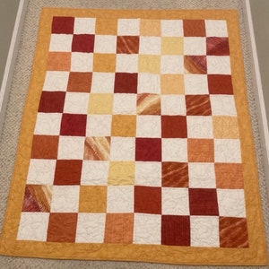 May include: A handmade quilt with a checkerboard pattern. The squares alternate between white, various shades of brown, and yellow. The quilt is bordered with a solid, golden-yellow fabric. The quilt is likely made of cotton.