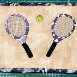 Könnte beinhalten: Ein Tischset im Tennis-Design mit zwei Tennisschlägern, lila und weißen Tupfen und schwarzen Griffen. Zwischen den Schlägern liegt ein grüner Tennisball. Das Tischset hat einen beigefarbenen Hintergrund und einen blau-weiß gemusterten Rand.