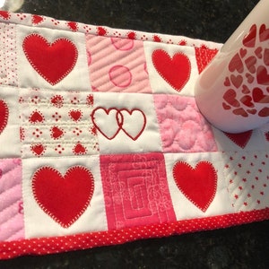 May include: A red and white quilted mug rug with a patchwork design featuring hearts. The mug rug is made of fabric squares with different patterns, including polka dots, stripes, and flowers. The mug rug is perfect for protecting your table from hot mugs.