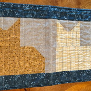 May include: A quilted table runner with a blue and white speckled border. The center features two cat-shaped patches, one in brown and gold and the other in yellow and white.