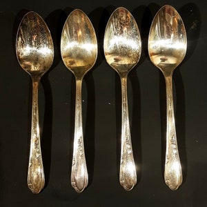 May include: Four silver spoons with ornate designs on the handles. The spoons are arranged in a row, with the handles pointing towards the viewer.