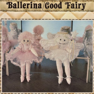 May include: Two handmade fabric dolls dressed as a ballerina and a fairy. The ballerina doll is wearing a pink tutu and has a pink curly wig. The fairy doll is wearing a white tutu and has a white curly wig. Both dolls are sitting on a wooden bar.