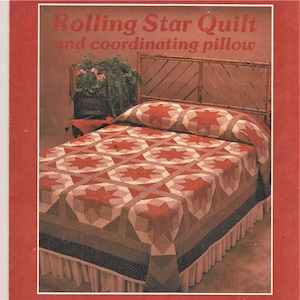 Op de afbeelding: Een rood-witte quilt met een sterpatroon, op een bed met een houten hoofdeinde. De quilt heet "Rolling Star Quilt and coordinating pillow".