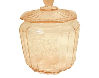 Pode incluir: Um pote de biscoitos vintage de vidro rosa com um design floral e uma tampa. O pote tem um design canelado e é feito de vidro transparente com um tom rosado.
