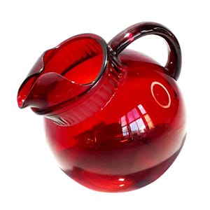 Puede incluir: Una jarra de vidrio rojo rubí con un cuerpo redondeado y un asa curva. La jarra tiene un pico para verter fácilmente. El vidrio es transparente, permitiendo el paso de la luz. Una pieza clásica para servir bebidas.