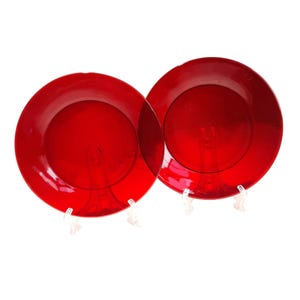 May include: Two red glass plates. The plates are round with a slightly raised edge. The plates are displayed on clear stands against a white background. The plates are a vibrant, solid red colour.
