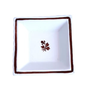 May include: Square white ceramic dish with a brown border and a floral design in the center. The dish has a slightly textured edge. The brown border frames the white interior, creating a simple yet elegant aesthetic.