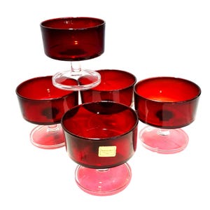 May include: A set of six ruby red glass dessert bowls. Each bowl has a short stem and a wide, shallow bowl. The bowls are arranged on a white background, with the light reflecting off the glass.