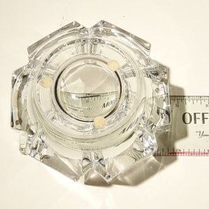 May include: A clear glass ashtray with a hexagonal shape and a round centre. The ashtray has three small white dots on the inside.