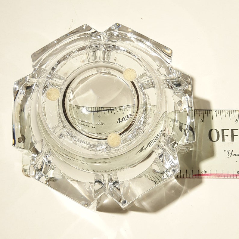 May include: A clear glass ashtray with a hexagonal shape and a round centre. The ashtray has three small white dots on the inside.