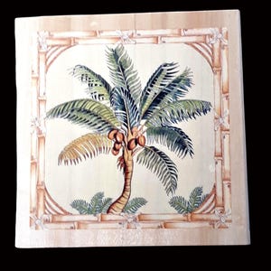 May include: A square, wooden decorative tile featuring a detailed watercolour painting of a palm tree with coconuts. The tree is surrounded by green foliage and framed by a bamboo border. The colour palette includes shades of green, brown, and beige.