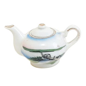 May include: A small, white porcelain teapot with a lid and handle. The teapot features a painted landscape scene with a boat, trees, and water. Gold trim accents the spout, handle, and lid. The teapot is a miniature size.