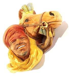 May include: A ceramic wall sculpture of a man wearing a red turban and a yellow scarf, with a horse's head peeking out from behind him.