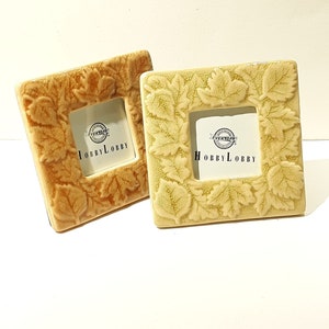 May include: Two ceramic picture frames with a leaf design. The frames are both square and have a small opening for a picture. The frames are both beige with a slightly different shade of beige. The frames both have the words "Everyday Hobby Lobby" printed on them.