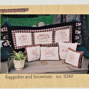 May include: A set of five embroidered pillow covers featuring rag dolls and snowmen with various sayings, including "Love Much", "Laugh Often", "Live Simply", and "Love Much, Laugh Often, Live Simply". The pillows are displayed on a wooden bench with a plaid fabric background.