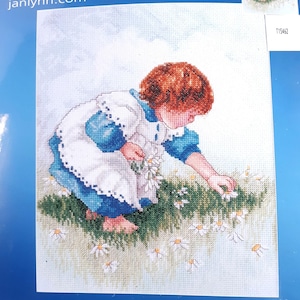 May include: A young girl with red hair wearing a blue and white dress picks daisies in a field.