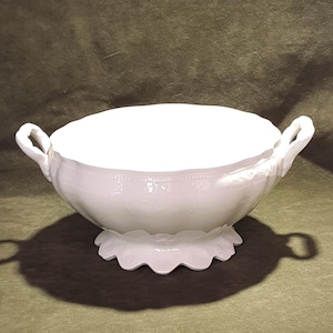 May include: A white ceramic serving bowl featuring two side handles, a scalloped base, and a subtle embossed beaded pattern along the upper rim. The bowl has a wide, open top with a smooth interior and a curved, rounded body shape.