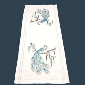 May include: A rectangular white table runner featuring two embroidered peacock motifs. Each peacock is perched on a brown branch with green hanging foliage. The embroidery uses shades of blue, orange, and green thread on a plain white fabric.