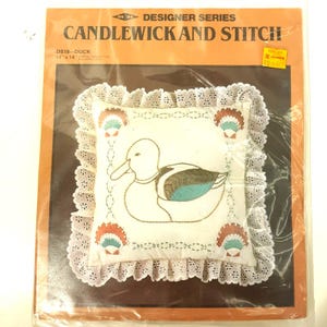 May include: A white pillow with a brown and blue embroidered duck design. The pillow is surrounded by a white lace trim. The package includes the pillow, embroidery floss, and instructions. The package is labeled "Candlewick and Stitch" and "DS18 - Duck".