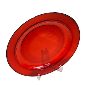May include: A striking red oval glass plate. The plate features a raised rim and a smooth, glossy finish. It is presented on a clear plastic stand, highlighting its elegant design. This decorative plate is suitable for serving or display.