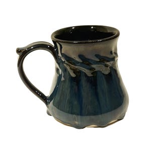 Puede incluir: Una taza de cerámica azul oscuro con un diseño único, hecho a mano. La taza tiene una base ancha y redondeada y un borde ligeramente acampanado. El asa está unida al lado de la taza y se curva hacia arriba.