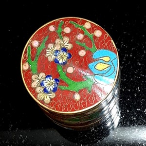 以下が含まれることがあります： 赤色の丸い小さな箱で、金色の縁取りと花柄のデザインが施されています。箱は青、緑、白、黄色のエナメルで装飾されています。蓋には青い花と緑の葉がついた花柄のデザインが施されています。