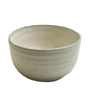 May include: A handcrafted, off-white ceramic bowl with a textured, ridged exterior. The bowl has a wide, rounded shape and a smooth interior. The rim is slightly uneven, adding to its rustic charm. Suitable for serving soups, cereals, or other dishes.