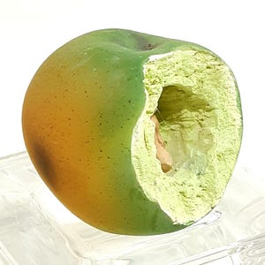 May include: A green and yellow apple with a bite taken out, revealing a light green interior and a small seed. The apple is on a clear, textured surface. The apple's skin transitions from green to yellow.