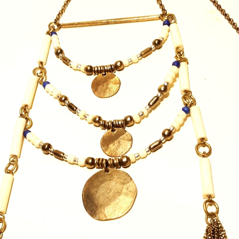 May include: A gold necklace with three layers of white and blue beads, and three gold circular pendants. The necklace has a gold bar at the top.
